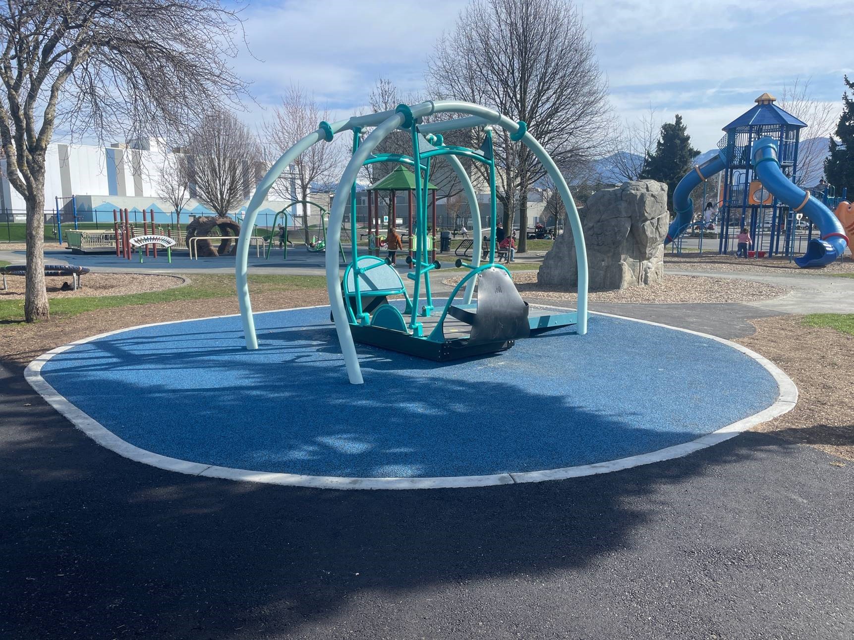 A blue and grey We-Go-Swing and a blue accessible rubber play surface. There are other playground structures in the background.